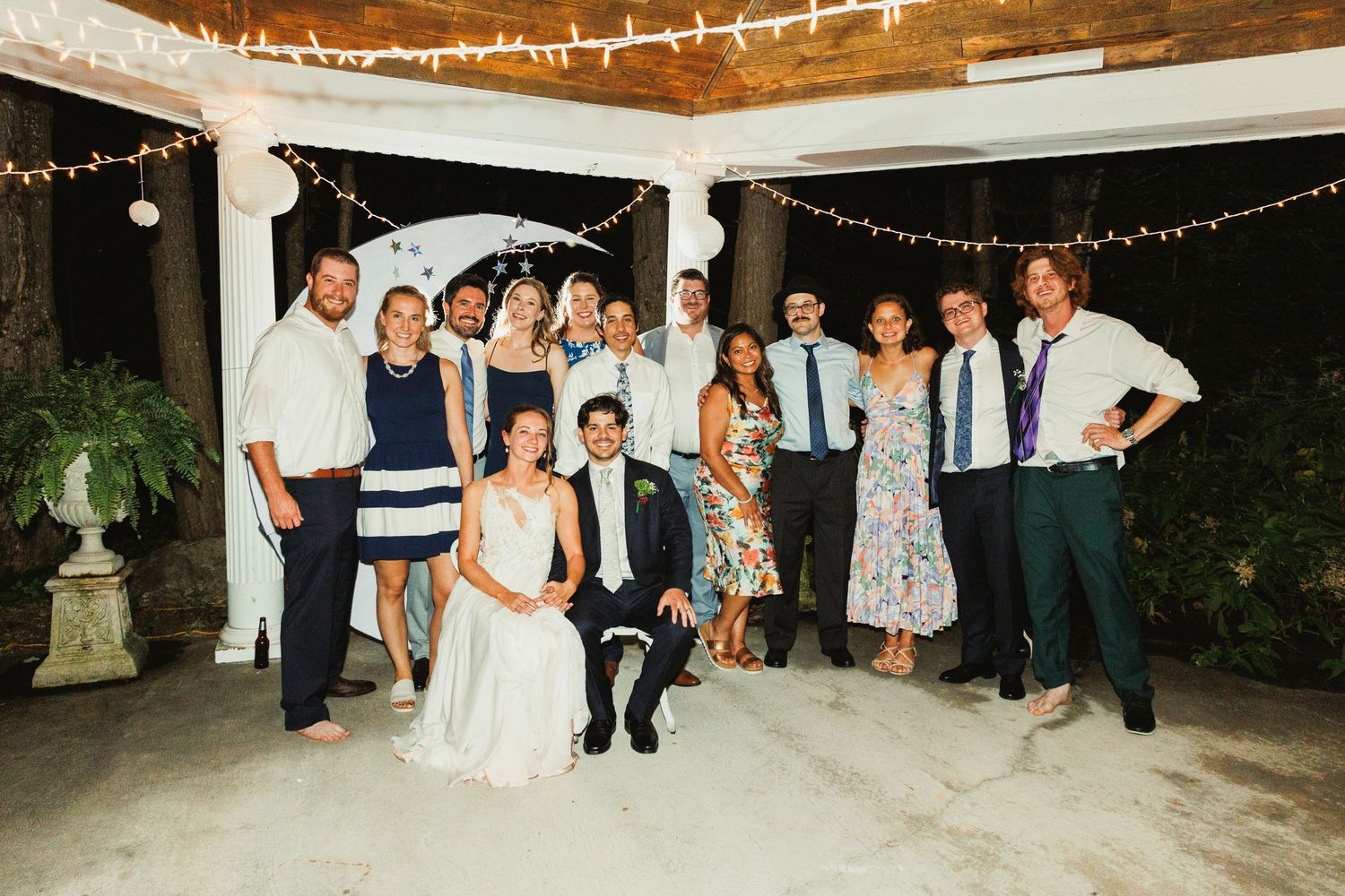Group photo of wedding guests under string lights at outdoor venue.