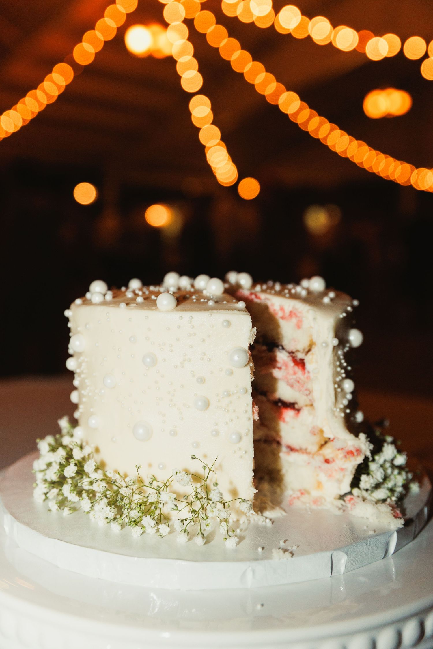 White wedding cake decorated with delicate baby's breath flowers and warm string lights in the background.