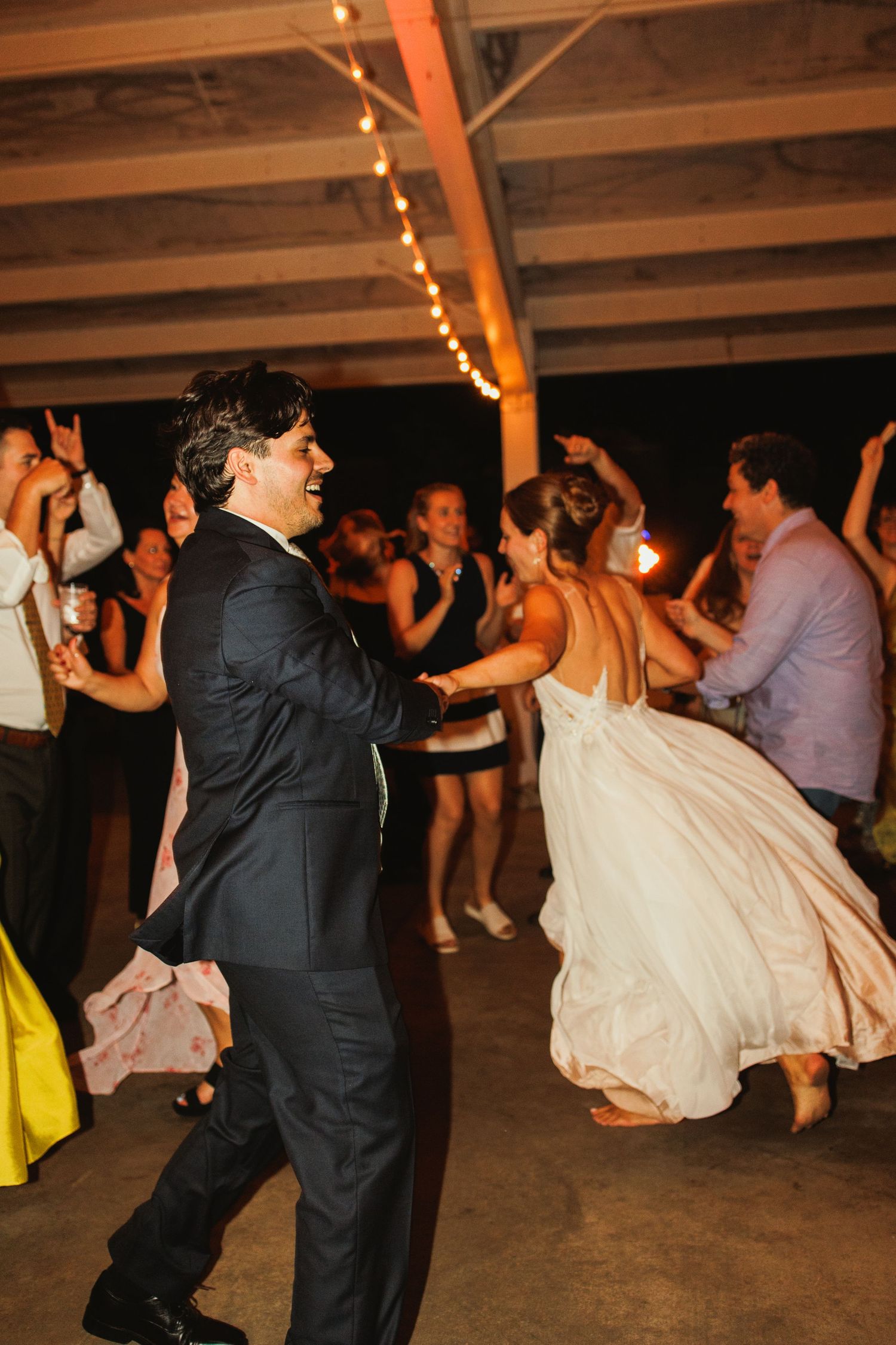 Wedding guests dancing energetically on a rustic indoor reception dance floor.