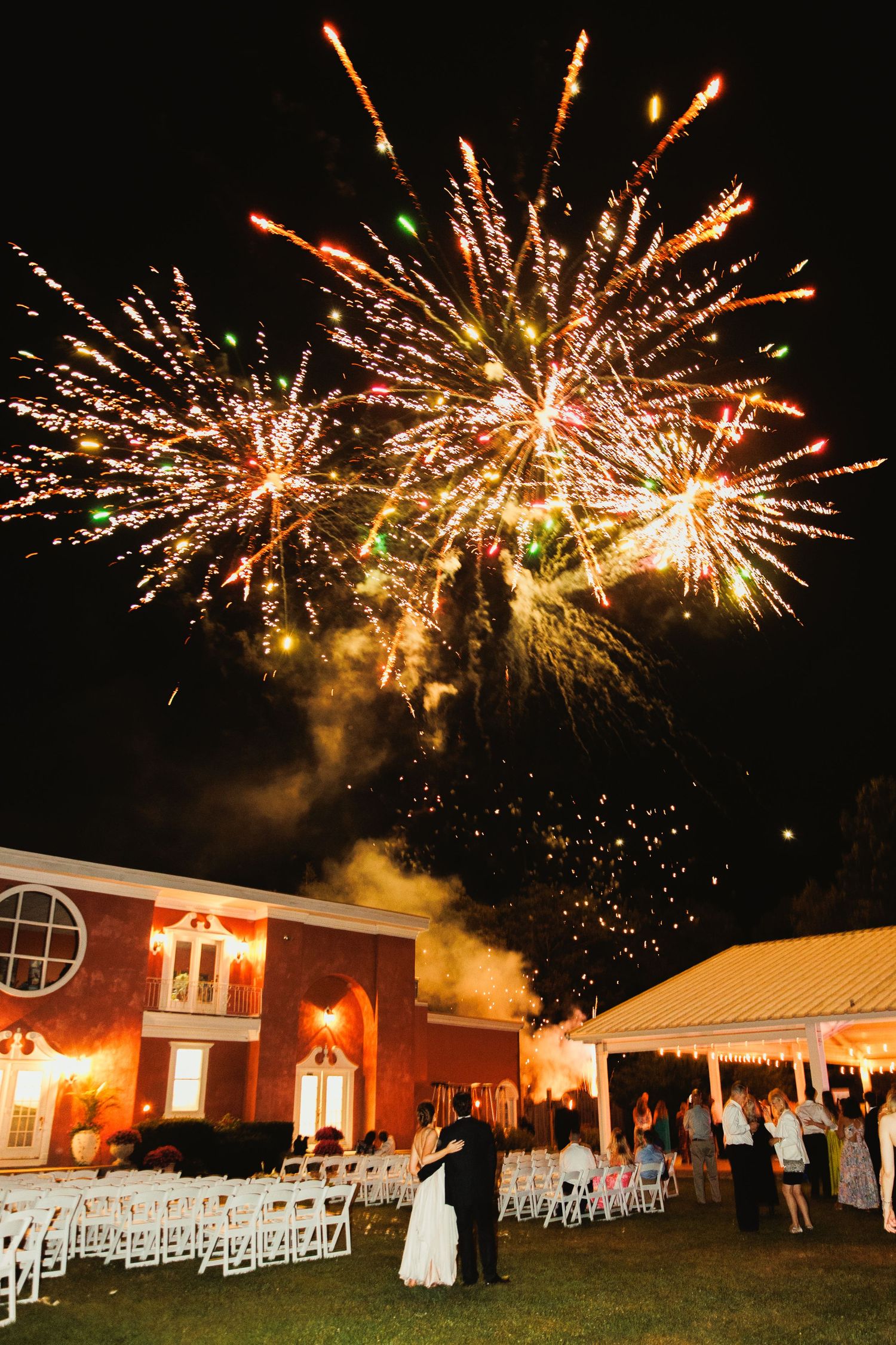 Spectacular fireworks display over a wedding venue with white chairs and brick building.