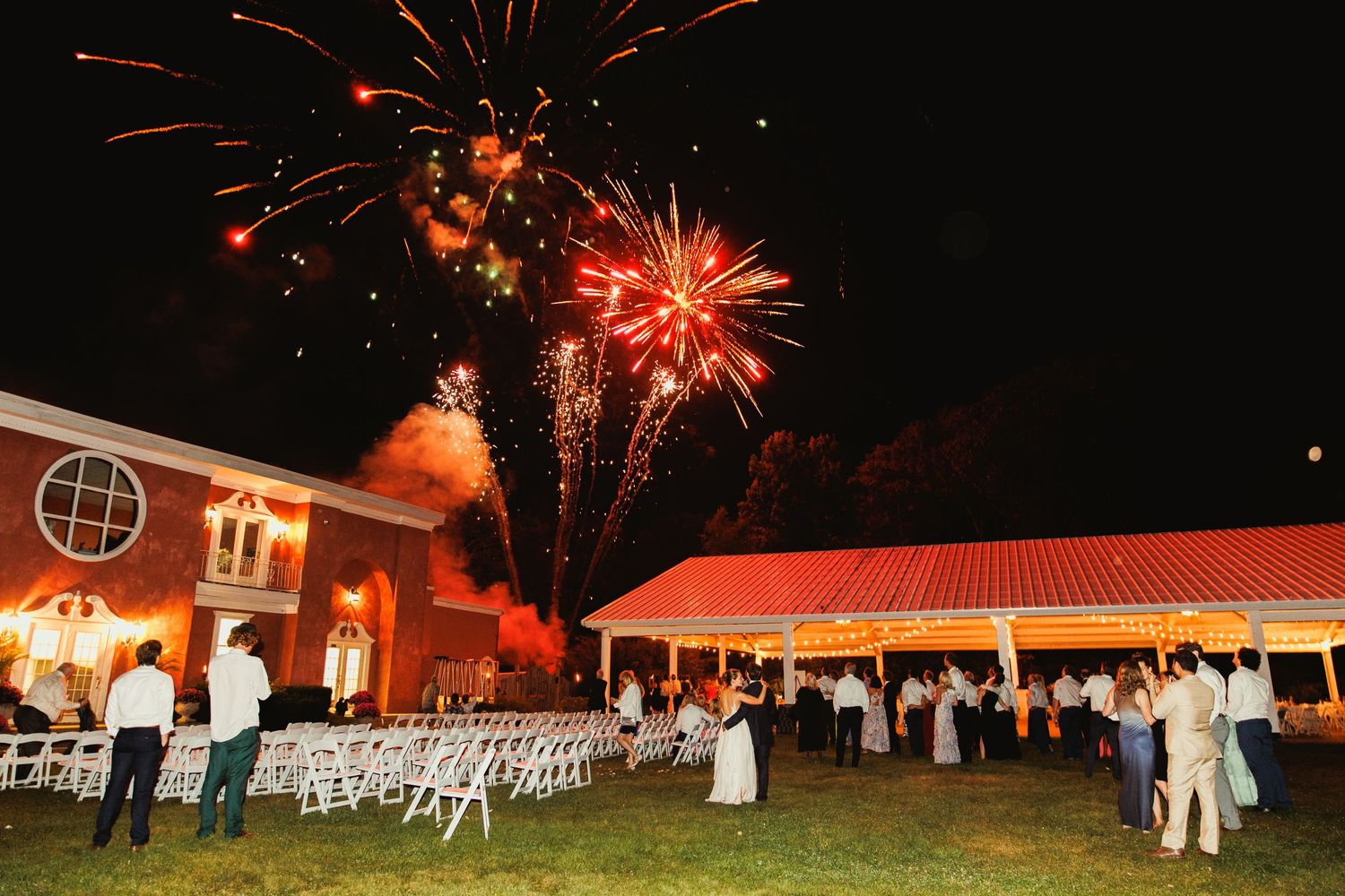 Fireworks display illuminating the night sky over an outdoor event venue with white chairs and tents.