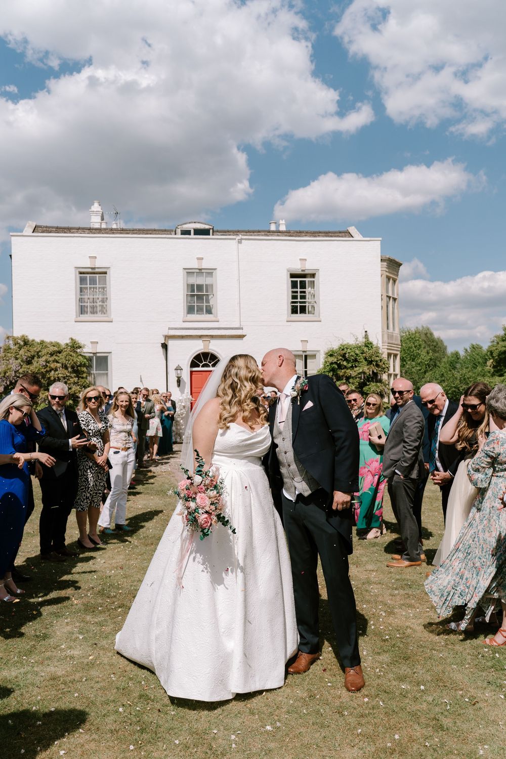 Wedding ceremony guests line up outdoors on a grassy hill next to a white building on a sunny day.