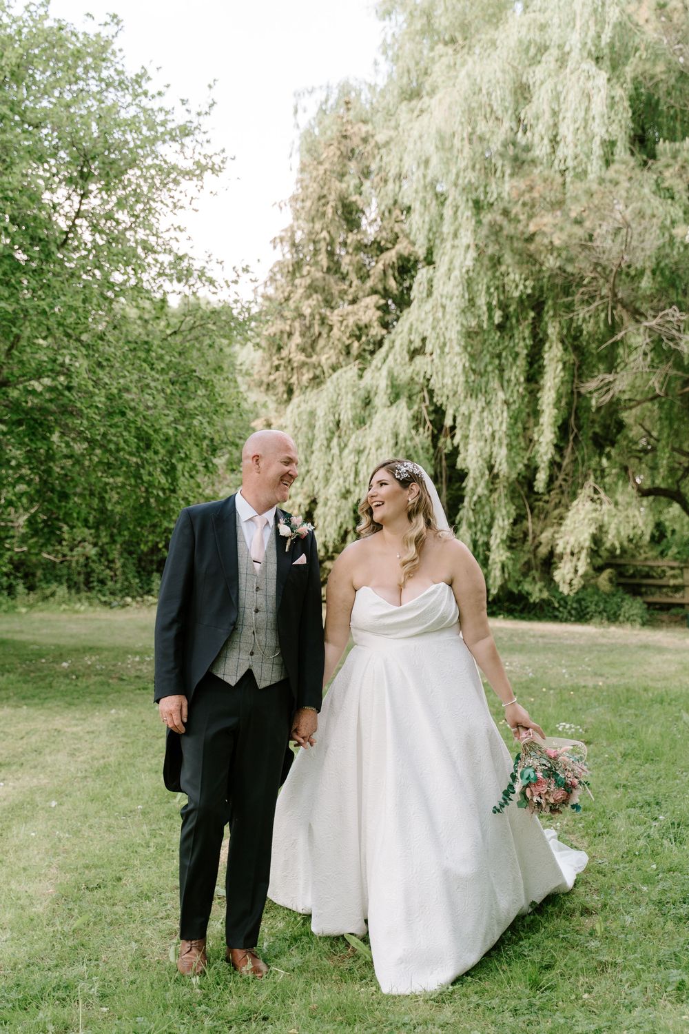 A couple in wedding attire shares a joyful moment under a weeping willow tree during their outdoor celebration.