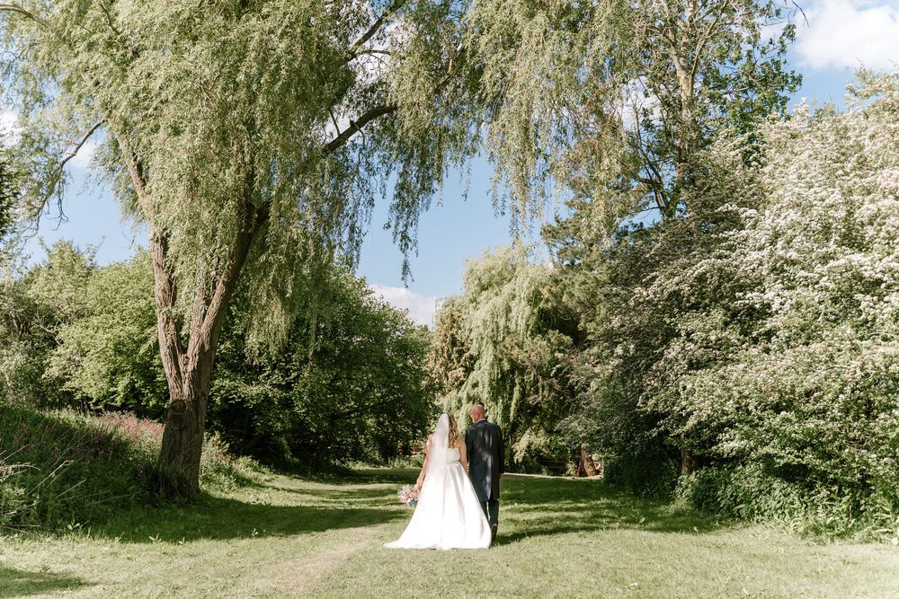 A romantic Worcestershire wedding couple stands beneath blooming willow trees in a lush garden setting on a sunny day.