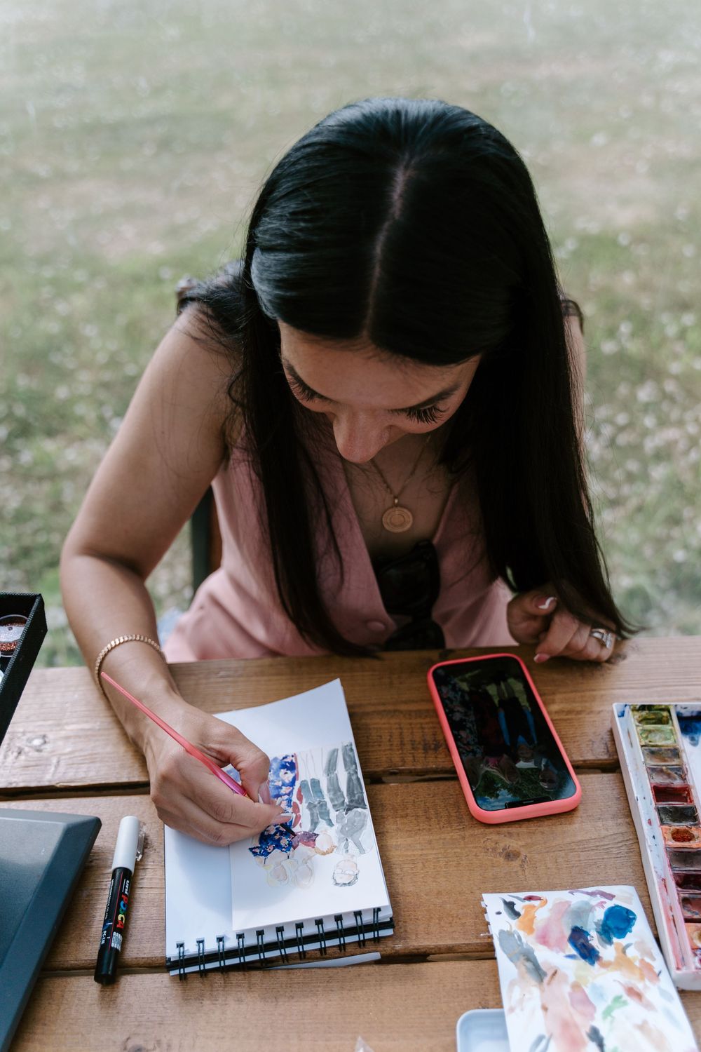 Worcestershire Artist sketching and painting in a sketchbook at a wooden table with art supplies and a phone nearby.