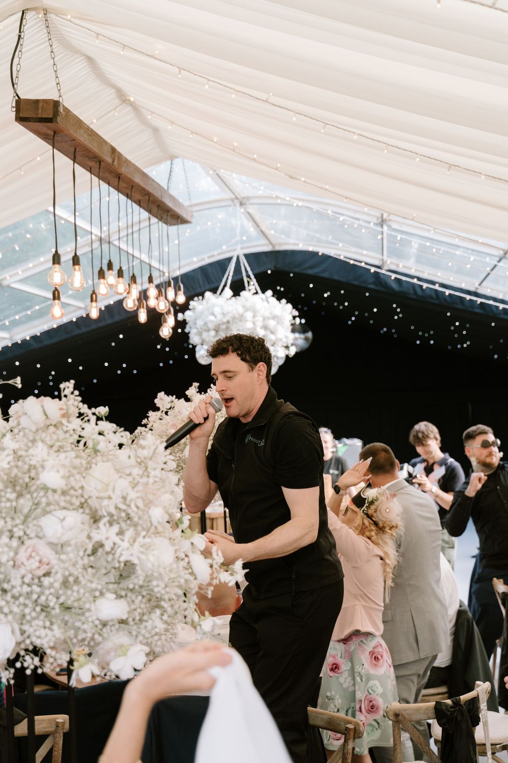 A wedding reception celebration under a white tent with elegant floral arrangements and hanging pendant lights.