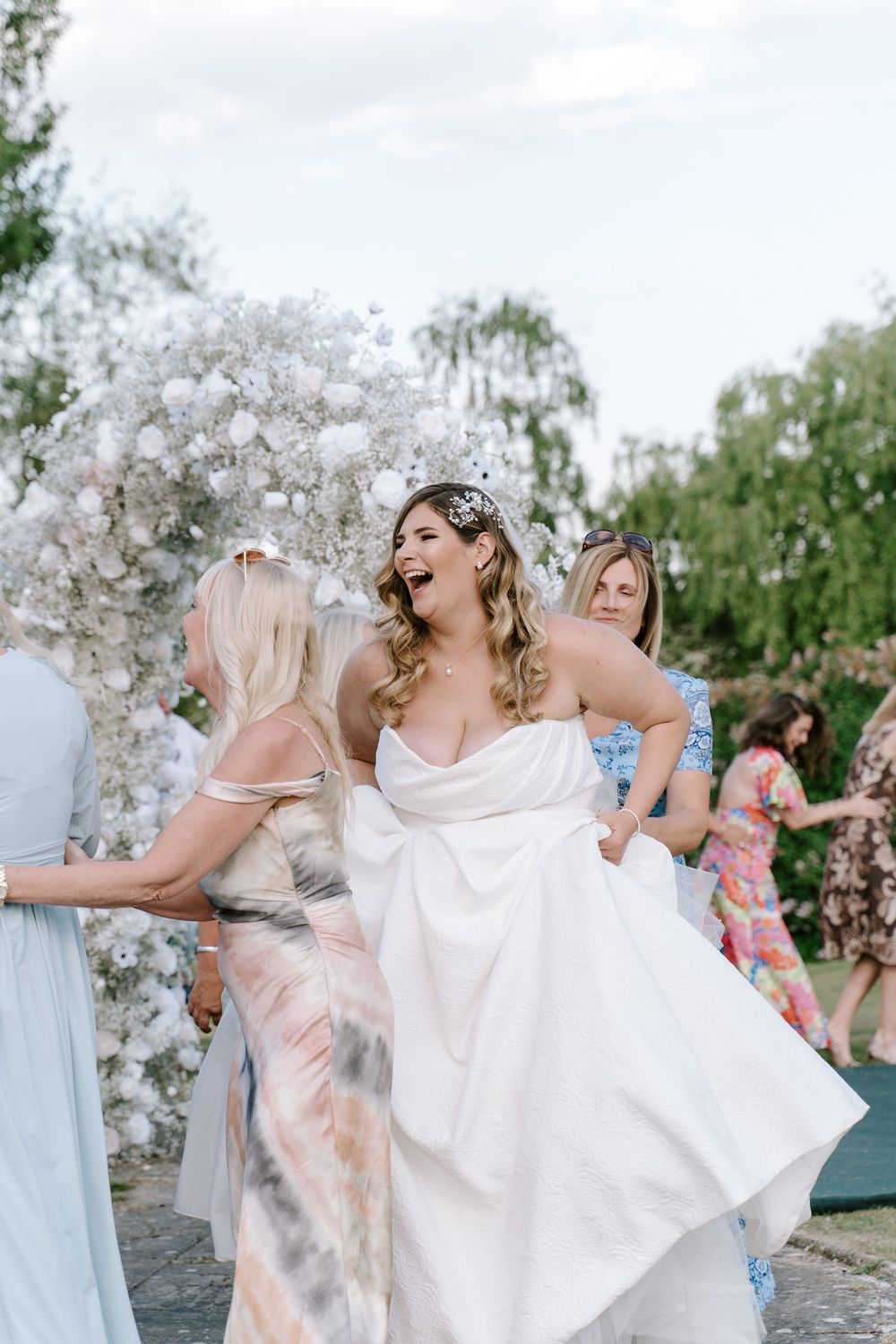 Cotswold Wedding party celebrates outdoors near white floral arch decorations in a garden setting.