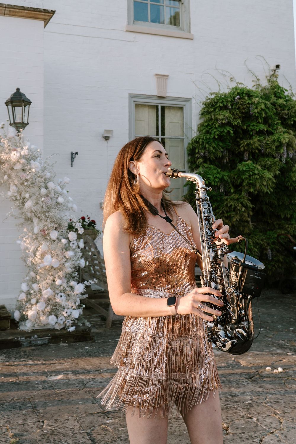 A Worcestershire musician in a sparkly dress performs with a saxophone against a white building backdrop with climbing vines.