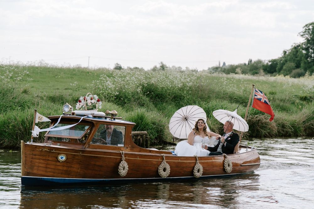 Wedding party celebrates on a classic wooden boat cruising down a river with white parasols at a Worcester wedding venue