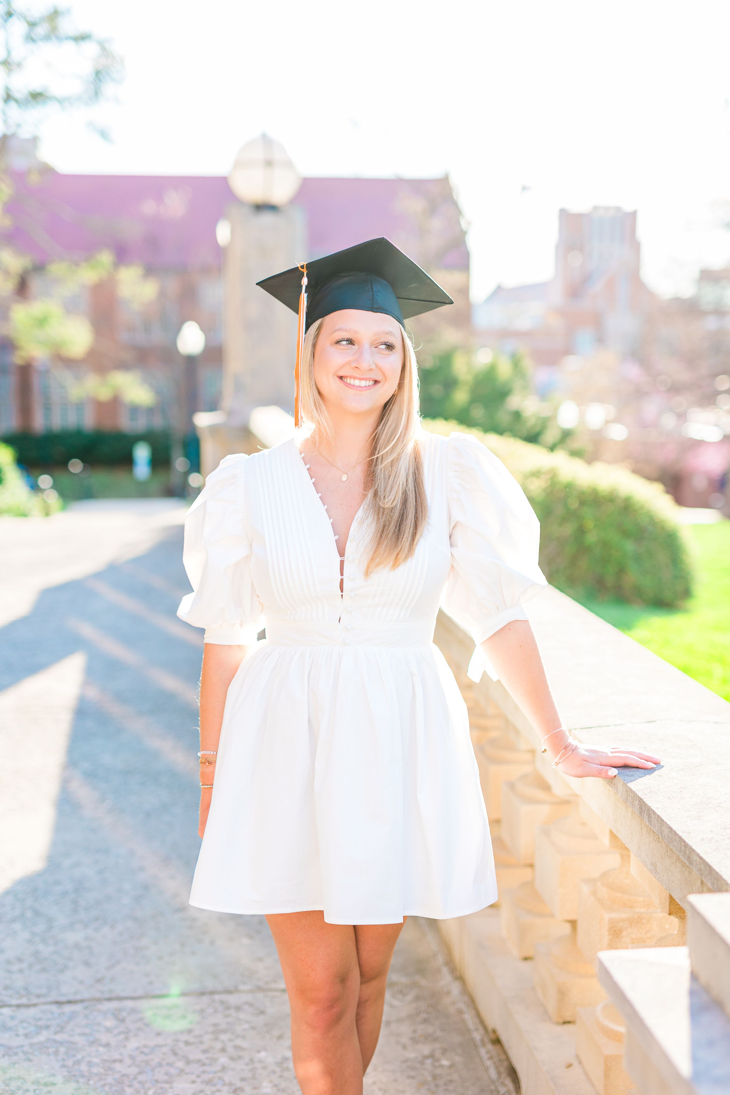 Graduate in white dress and cap poses outdoors on sunny day for graduation photos.