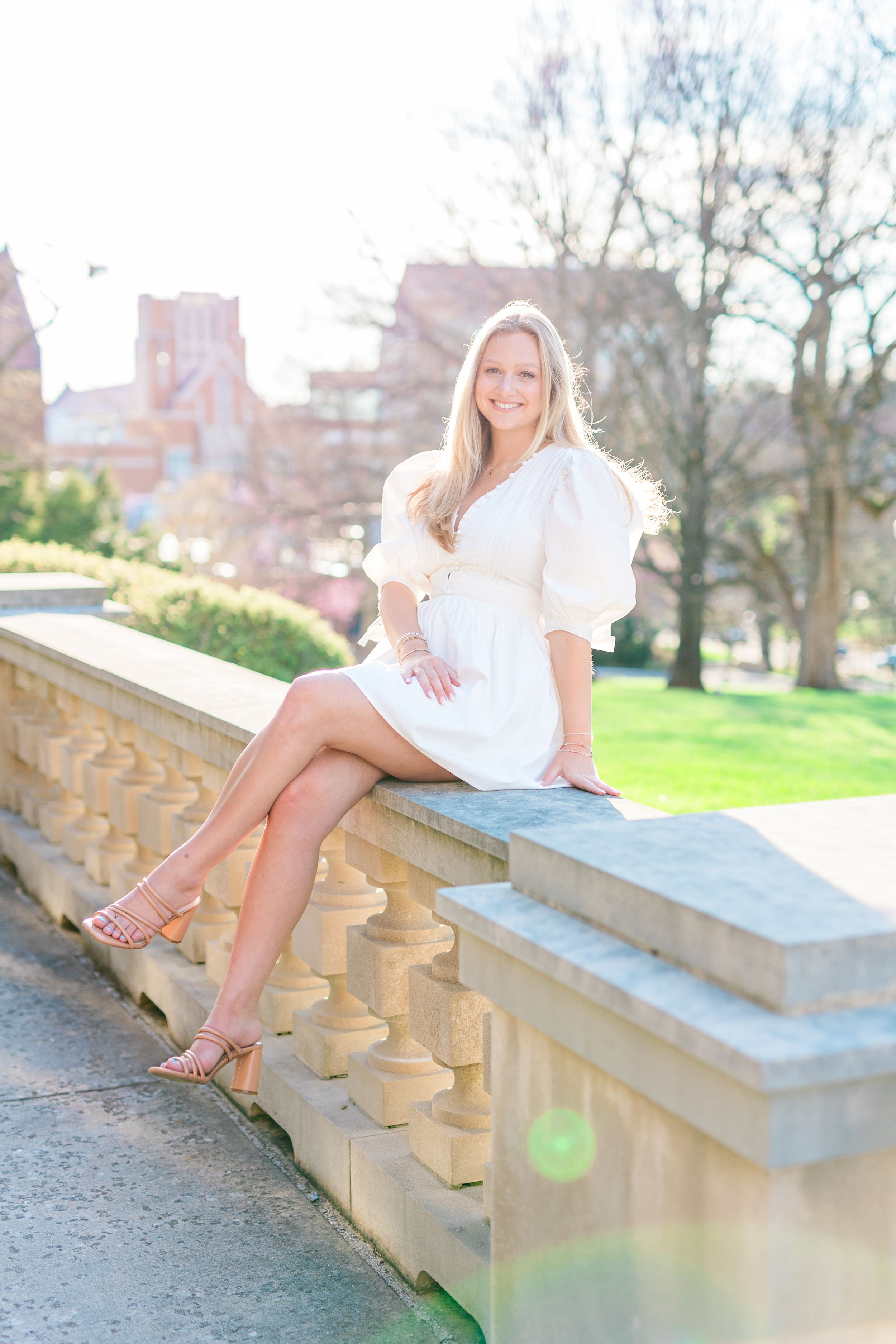 Woman in white dress sitting on stone wall with city buildings and green lawn in background on sunny spring day.