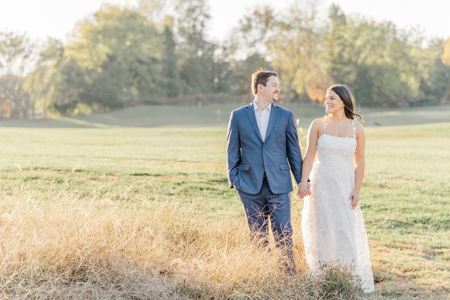 A couple in wedding attire walks through a golden field at sunset holding hands during their outdoor photo session.