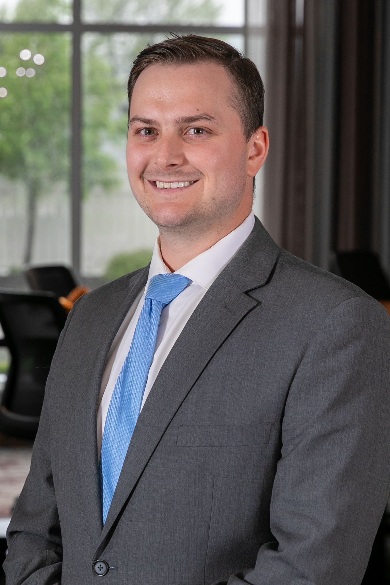 Professional headshot of a businessman in a grey suit and blue tie smiling in an office setting.