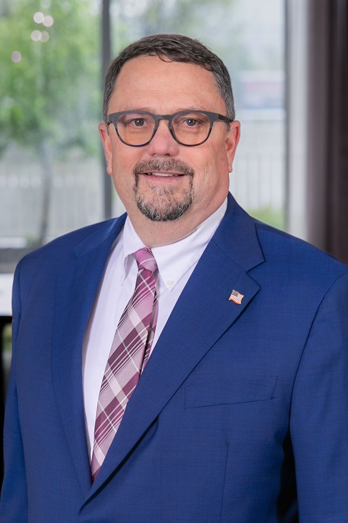 Professional headshot of someone in a navy suit with pink striped tie and glasses against blurred office background.