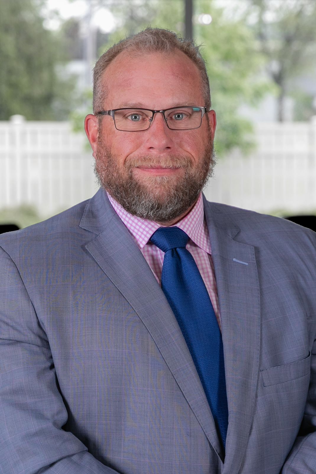 Professional headshot of someone in a gray suit with pink checkered shirt and blue tie against an outdoor background.