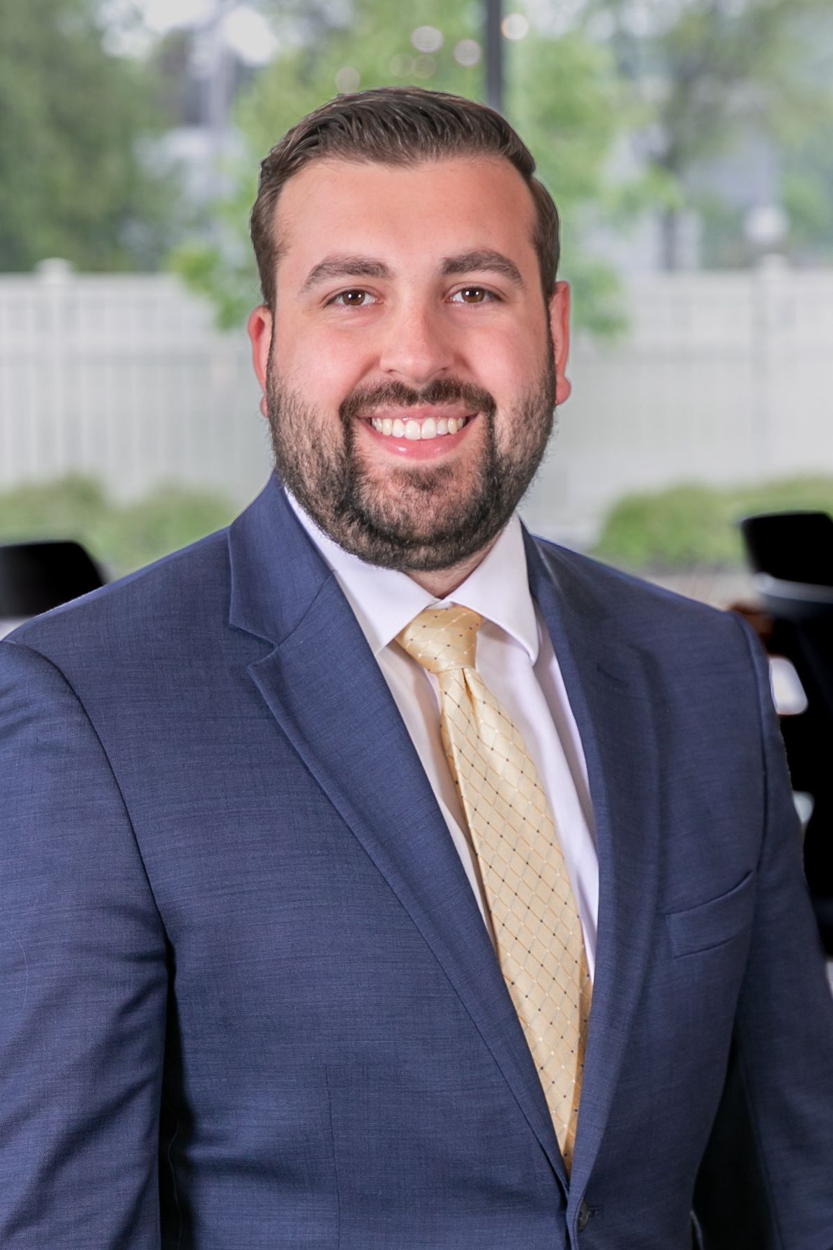 Professional headshot of a businessman wearing a navy suit and gold tie against a blurred office background.