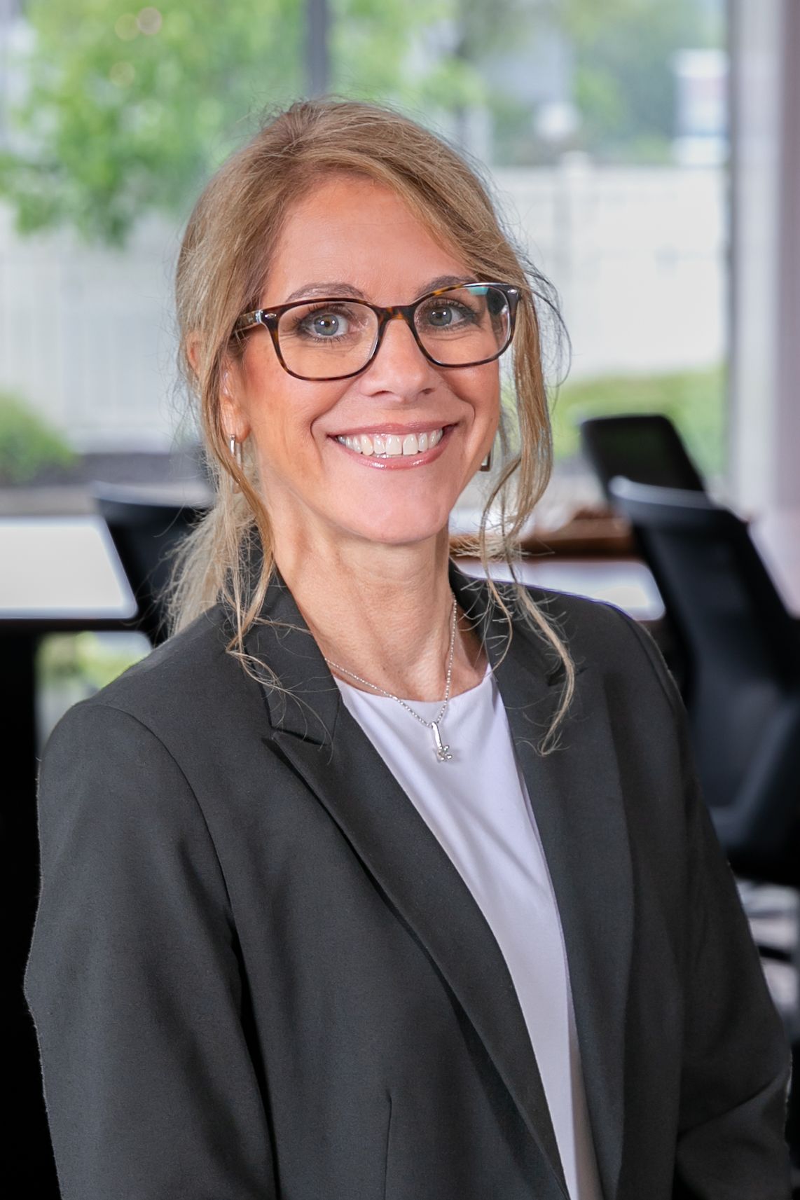 Professional wearing glasses and black blazer smiles warmly in modern office setting.