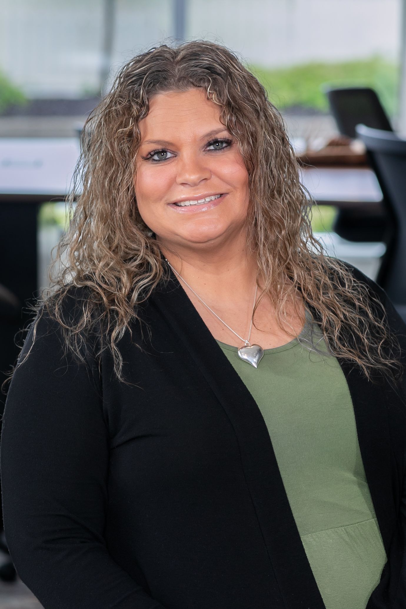 Professional headshot of someone in a black blazer and olive green top with curly hair smiling at the camera.