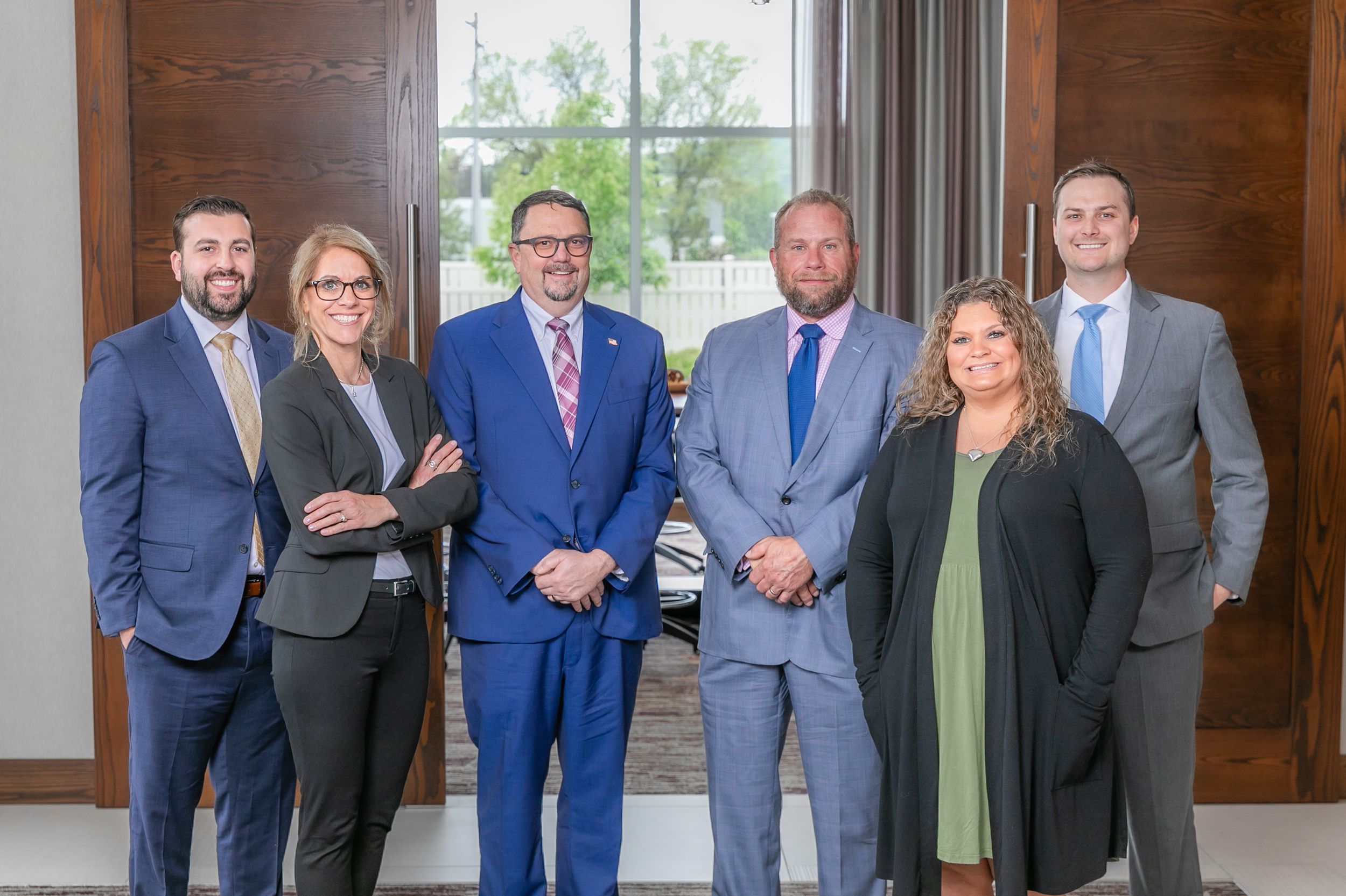 A group of professionals in business attire stand together against a wooden wall for a formal photo.