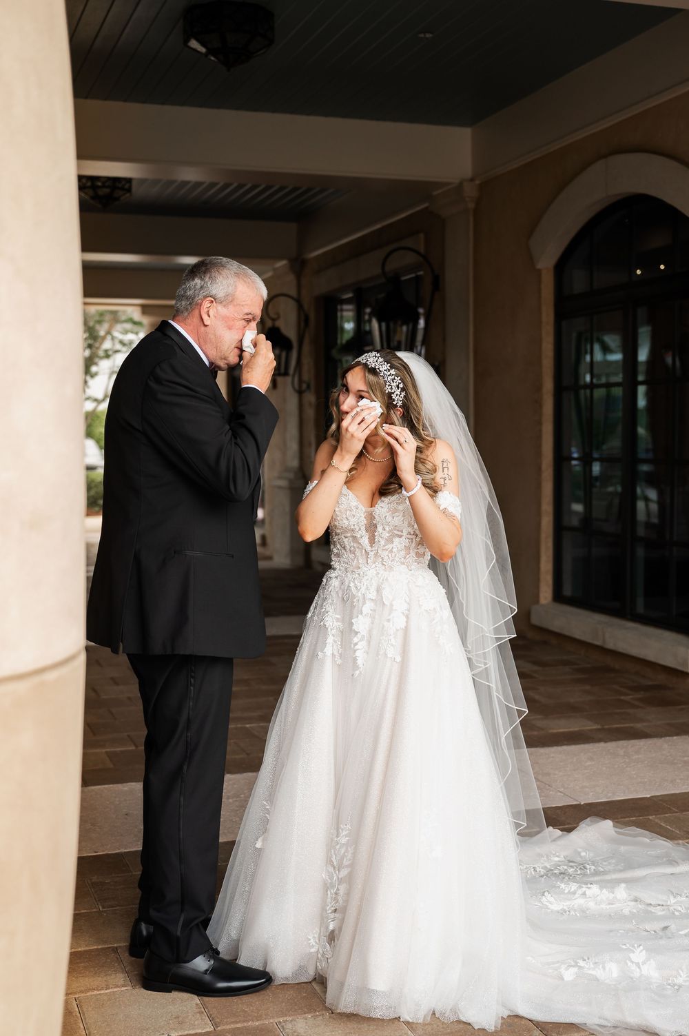 Emotional first look moment between bride in white gown and father in black suit under columned walkway.