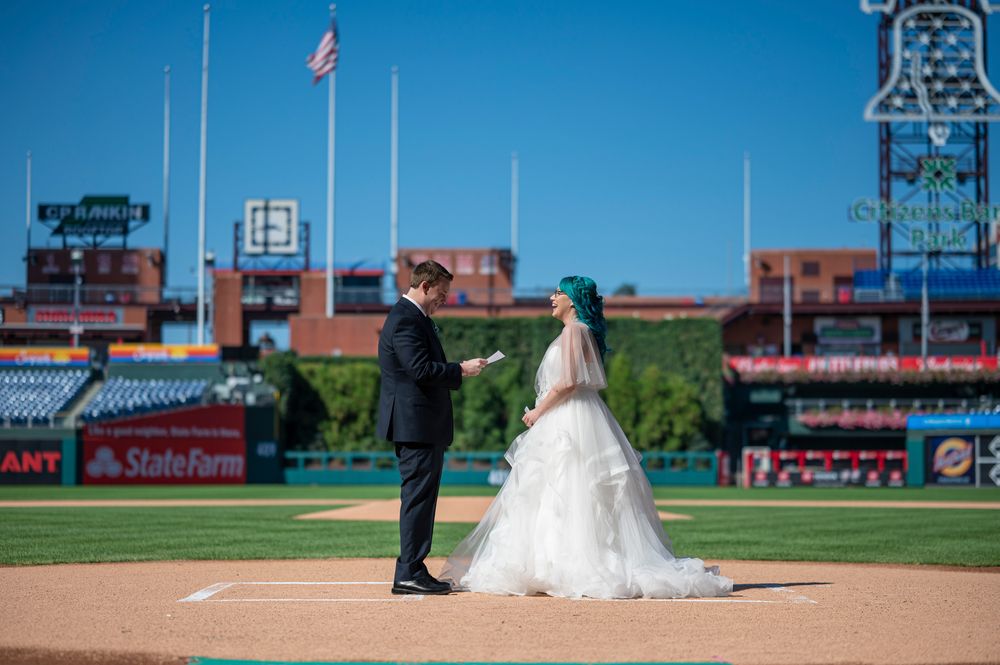 Wedding couple poses on baseball field with stadium seats and green grass visible in background.