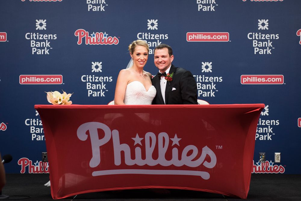 A red Phillies baseball team banner hangs against a backdrop of Citizens Bank Park signage and logos.
