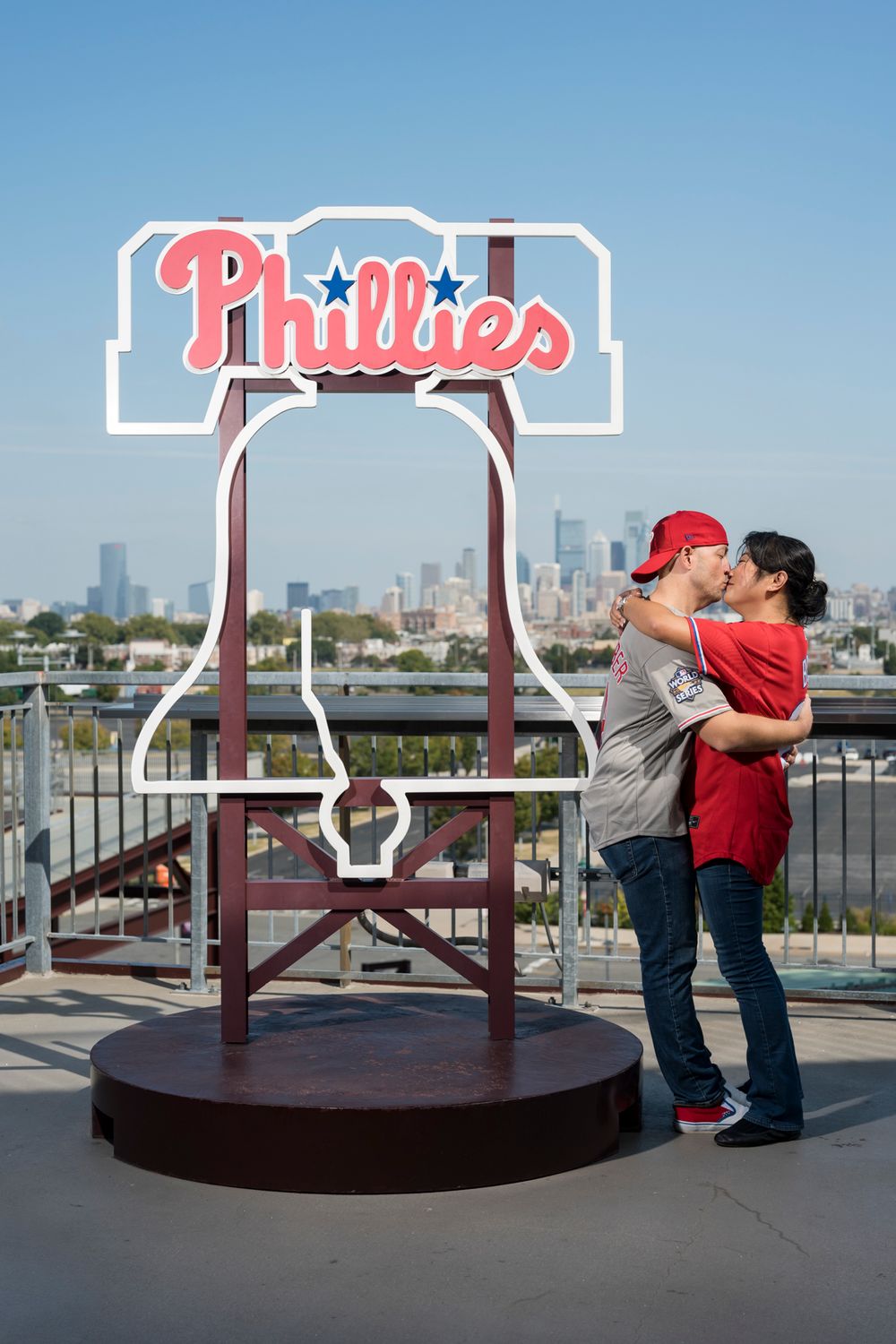A couple embraces in front of a large Philadelphia Phillies logo display with the city skyline visible in the background.