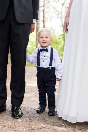 Liten gutt med sløye og bukseseler. Han smiler litt mot kamera. I bildet ser vi bare føttene til pappa og kjolen til mamma.
