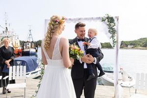 Bruden har akkurat kommet frem til blomsterbuen hvor brudgommen står med barnet på armen. Barnet ser opp  mot mamma´n sin.