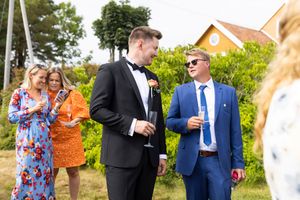 Øyeblikksbilde av brudgom og kompis. De ser mot hverandre. Bak dem står to andre gjester og ser på noe felles på en mobiltelefon.