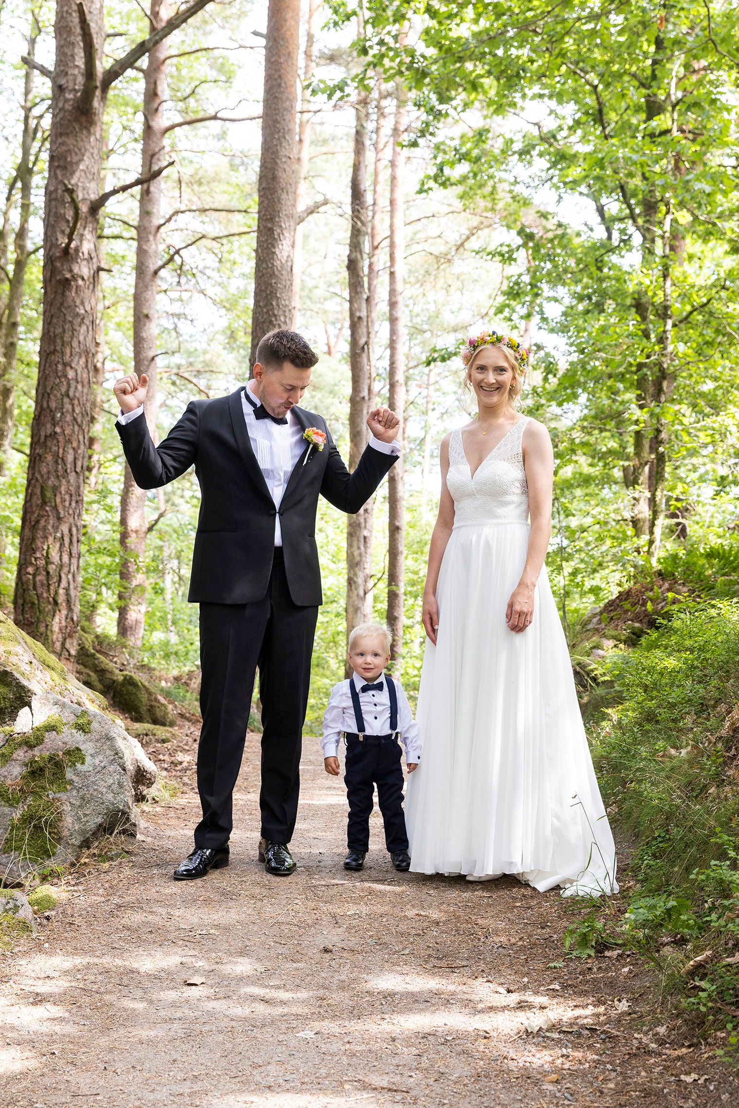 Familiebilde i skogen. Sønnen i bukseseler og sløye står på bakken. Bruden ser i kamera, mens brudgommen løfter hendene for å juble.