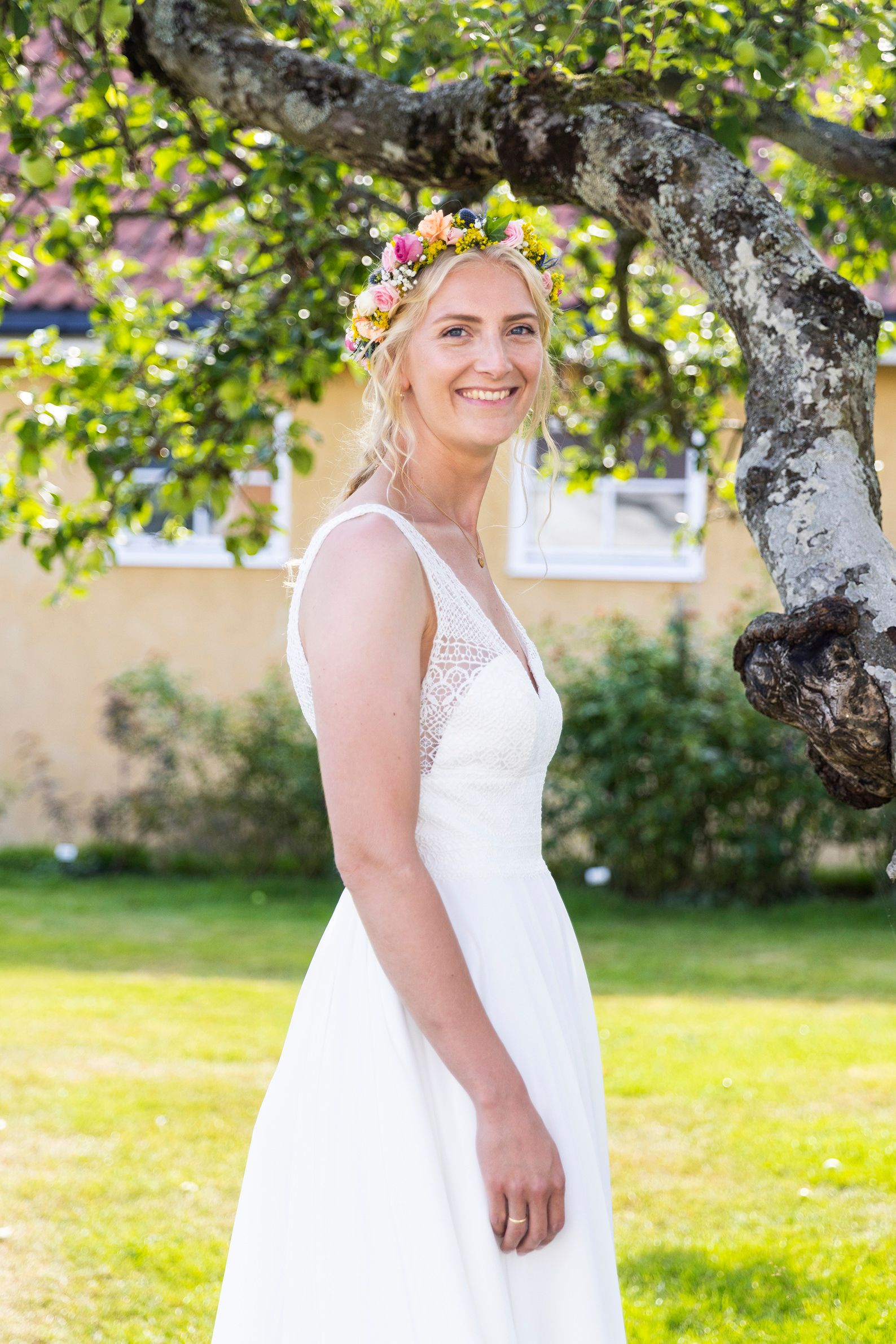 Bilde av bruden i hvit brudekjole og blomsterkrans i håret. Hun står foran et epletre i en nydelig hage.