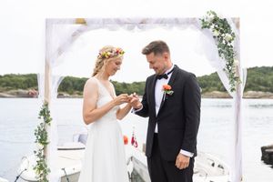 Nærbilde av brud som setter på ringen på mannen sin. De står foran en blomsterbue, bak dem ser vi havet.