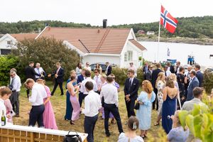 Oversiktsbilde av alle gjester som mingler i hagen. Vi ser det norske flagget er heist, havet og skjærgården i bakgrunnen.