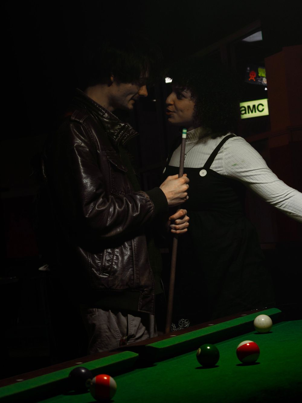 Players line up shots at a dimly lit pool table with green felt and red balls visible.