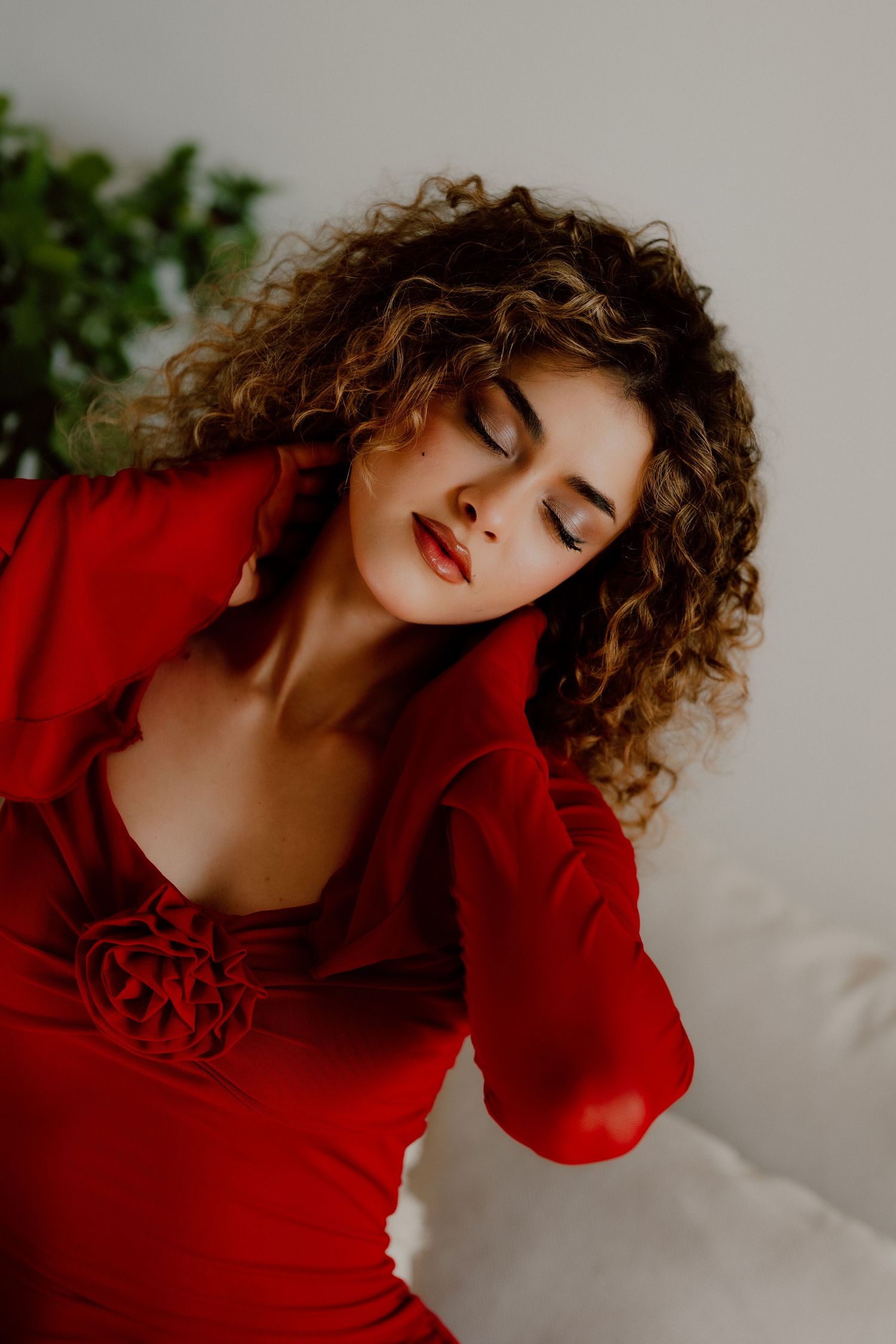Woman in red dress with curly hair and red lips poses gracefully with eyes closed in a serene indoor setting.