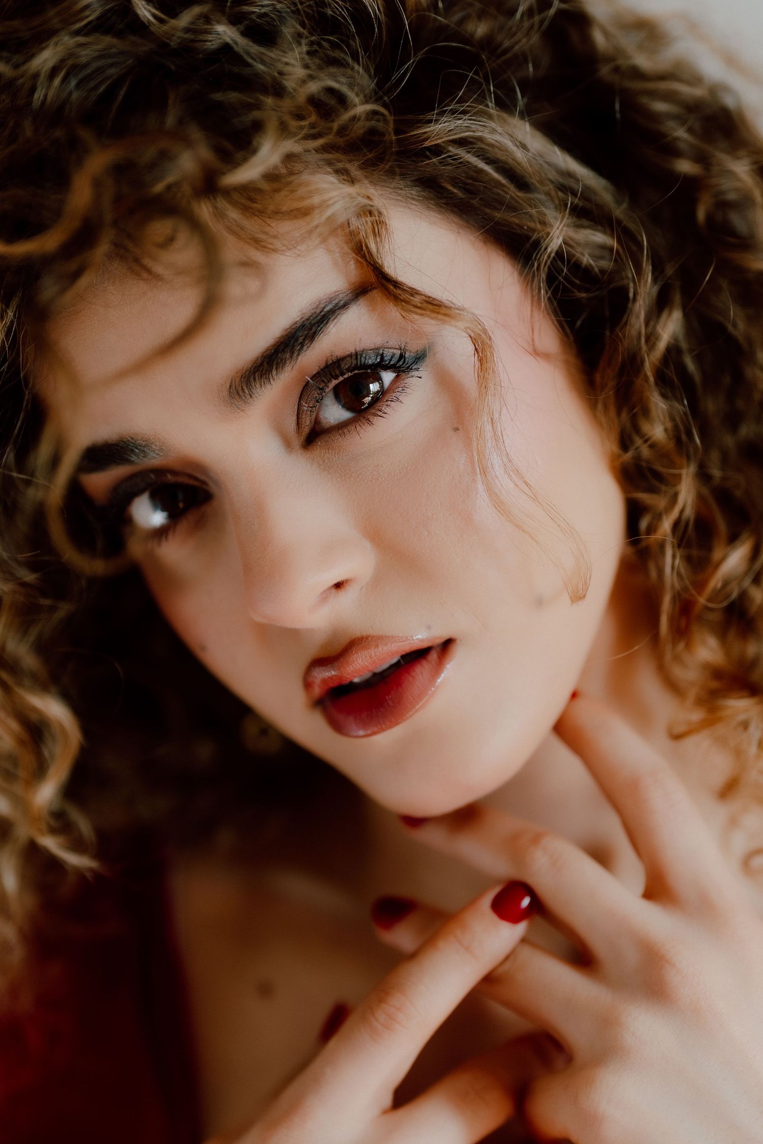 Close-up portrait of a young woman with curly hair, bold eye makeup, and red nails touching her neck.