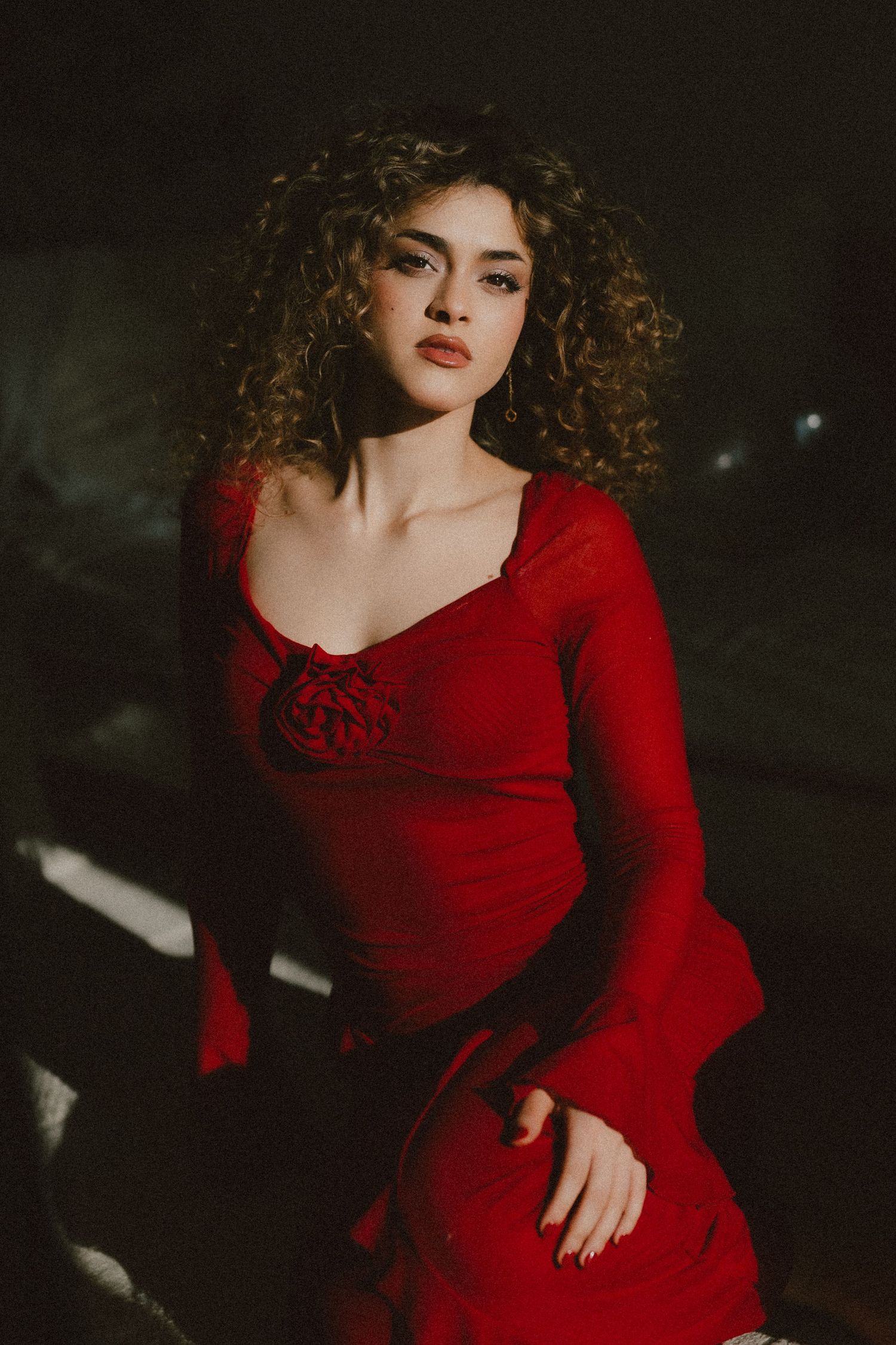 Young woman with curly hair poses in a red long-sleeve dress with floral detail in dramatic moody lighting.