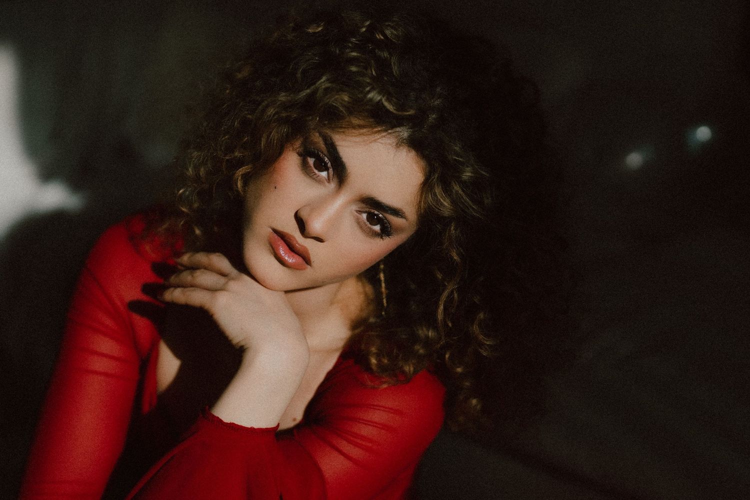 Young woman with curly dark hair in a red dress poses thoughtfully against a dark background.