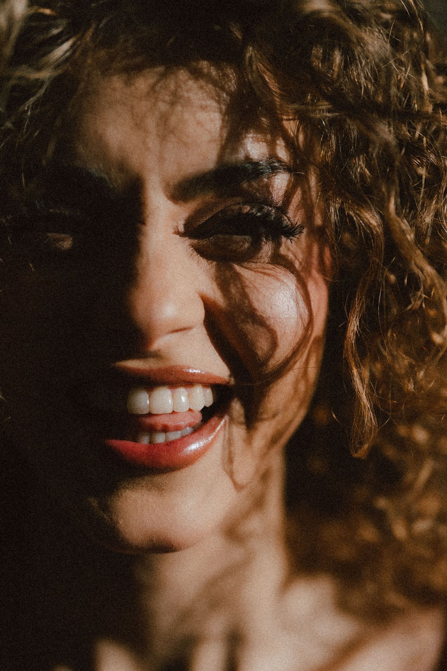 Close-up portrait of a smiling woman with curly hair and red lips in dramatic warm sunlight.