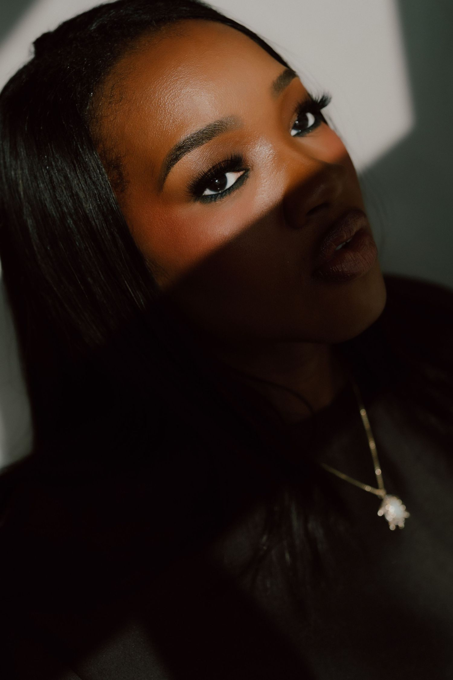 Close-up portrait of a  woman with sleek hair, gold necklace, and bold eye makeup in dramatic lighting.