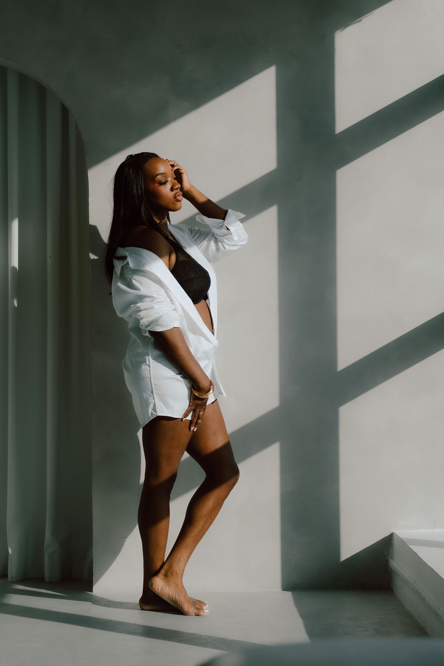 woman in a white open shirt and shorts leans against a sunlit wall, eyes closed, barefoot.