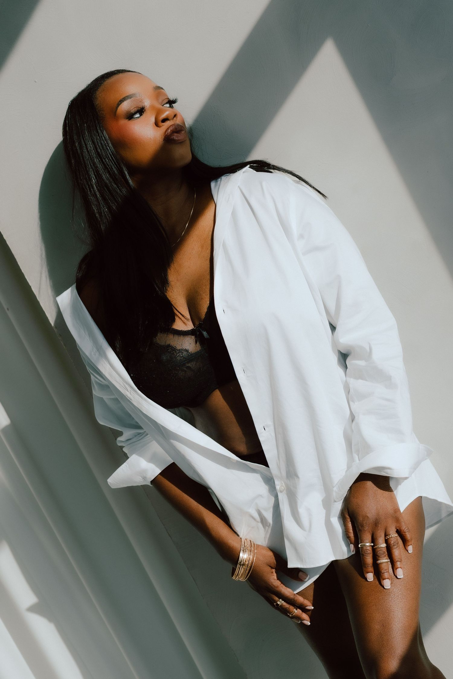 A confident woman poses in black lingerie and an open white oversized shirt, adorned with gold jewelry.