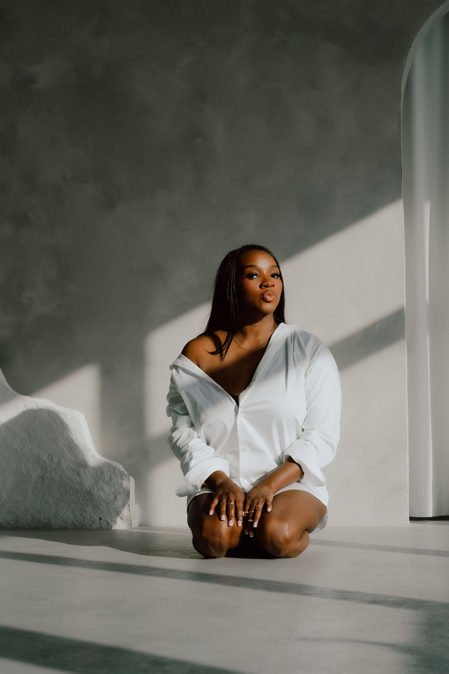A young woman in a white oversized shirt sits cross-legged on a studio floor bathed in natural light.
