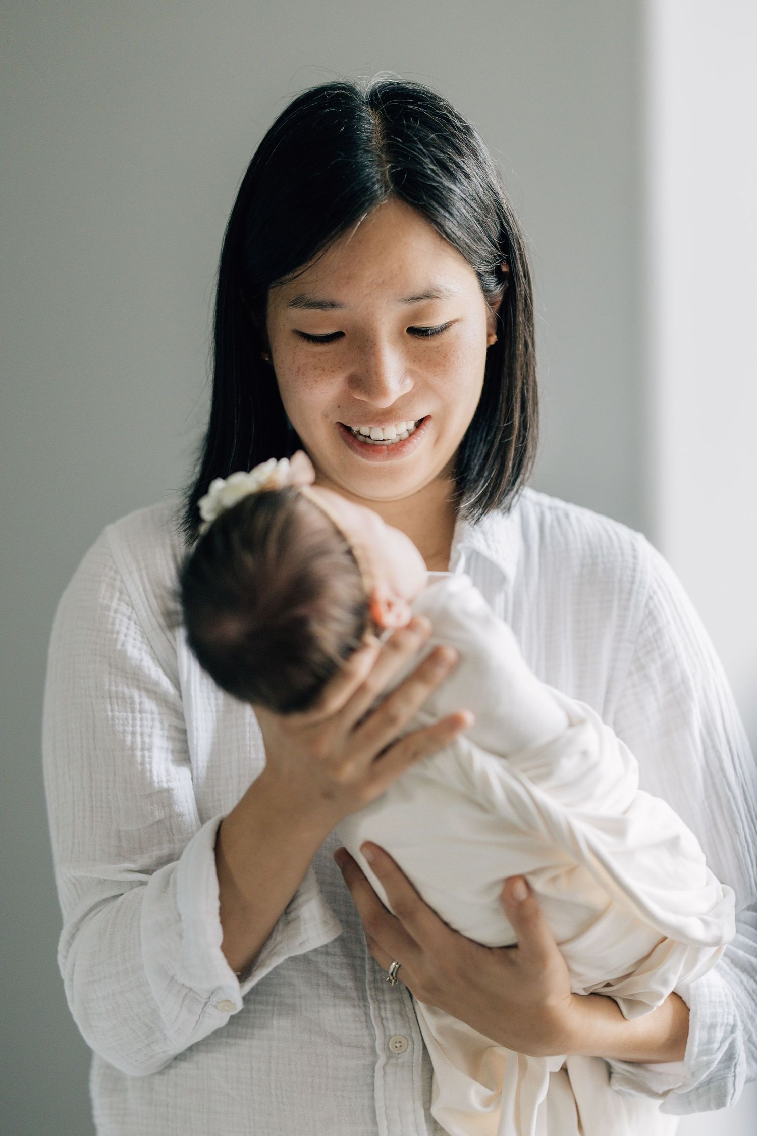 Smiling Asian mother in white shirt holding newborn baby girl with floral headband indoors.