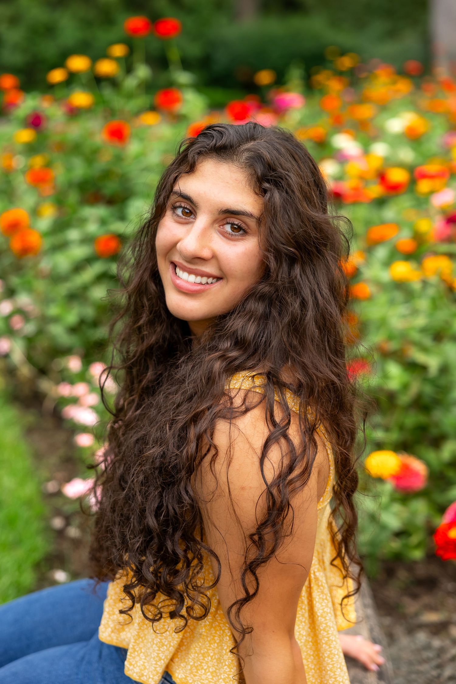 A person in a white floral dress poses playfully against lush green foliage in an outdoor setting.