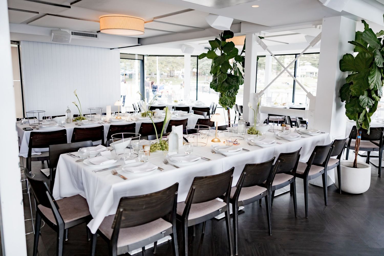 A modern restaurant interior with white tablecloths and place settings arranged in long rows under pendant lighting.