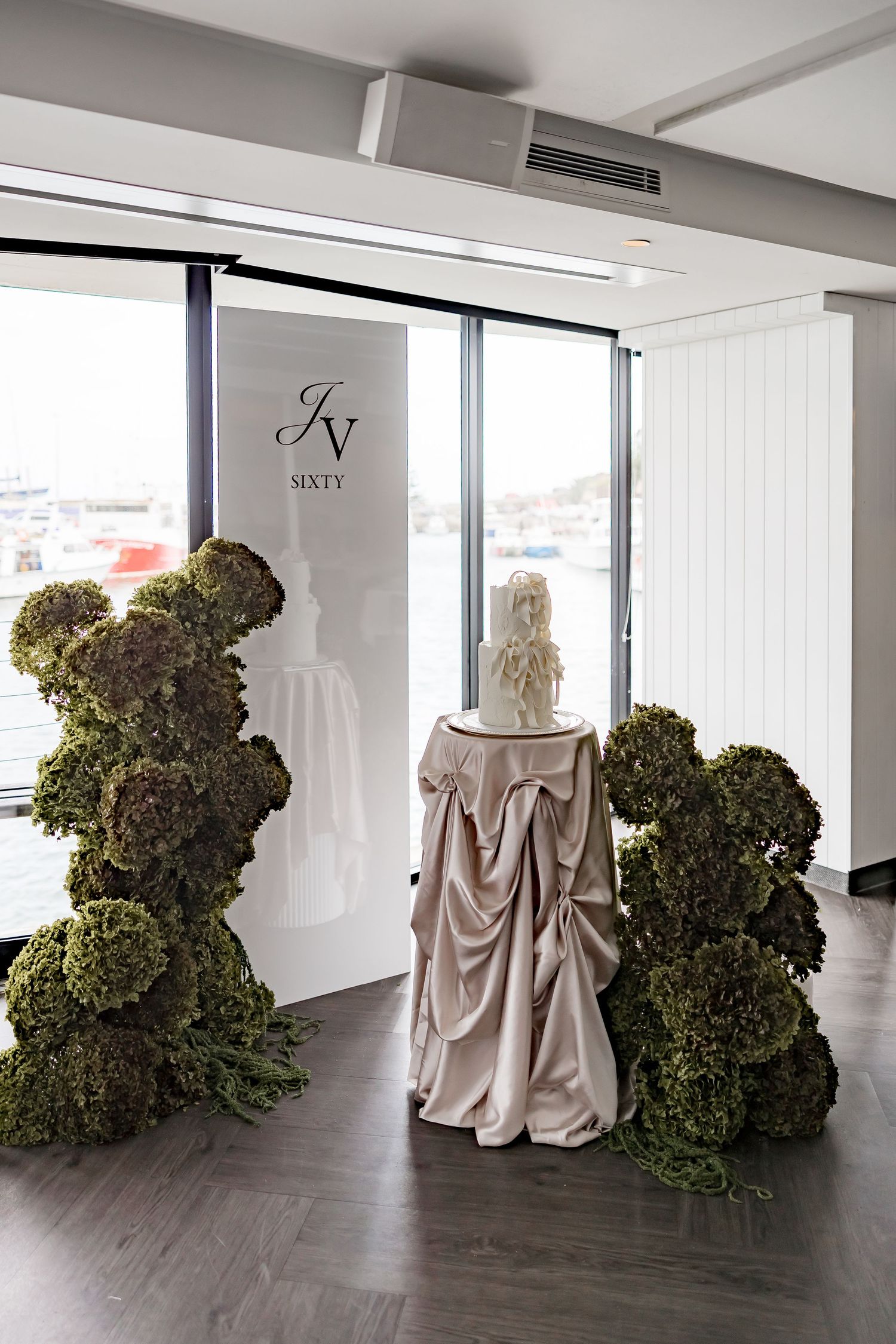 A series of mannequins draped in beige fabric stand beside large green moss-covered sculptural forms in a modern gallery space.