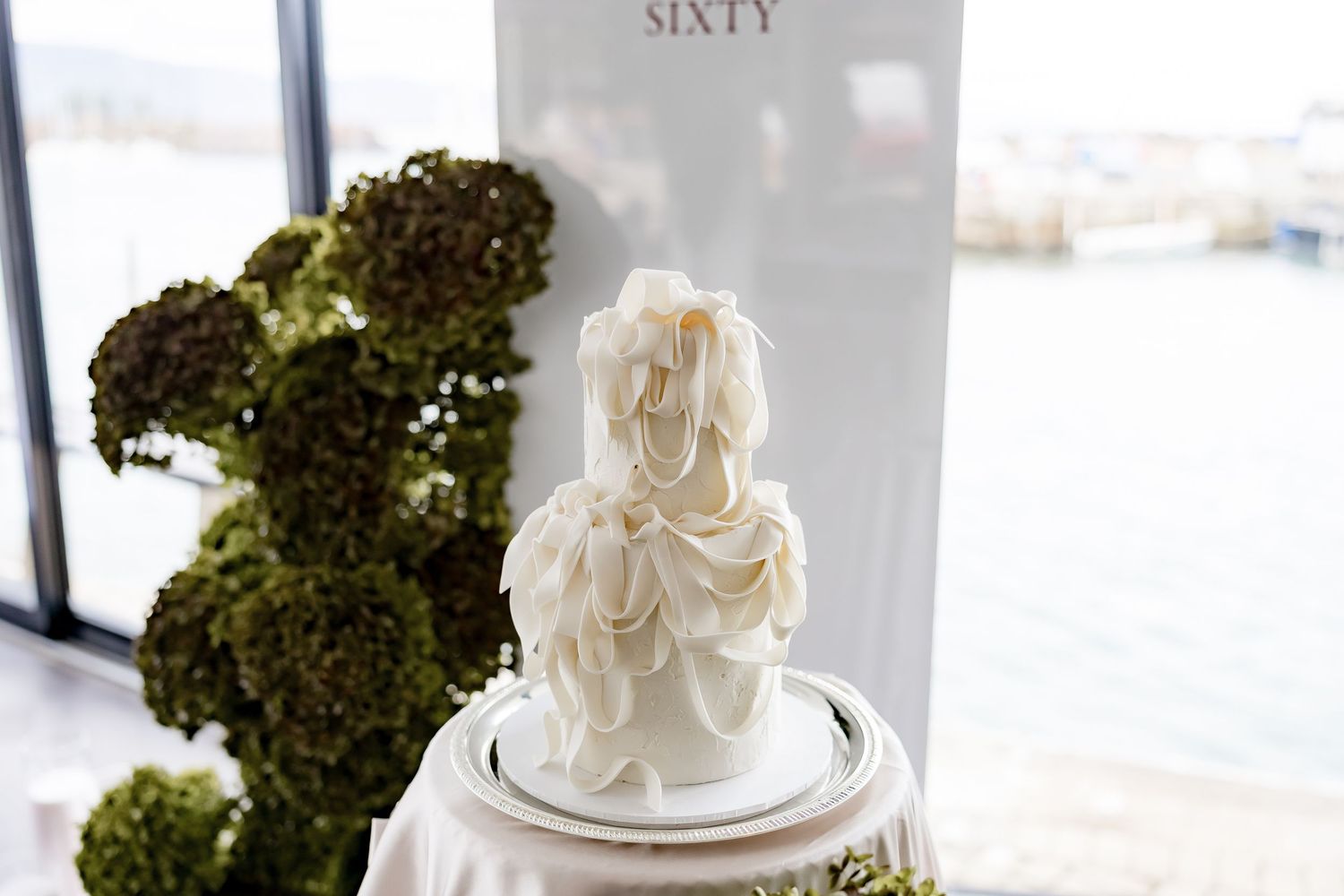 Elegant wedding display featuring white floral arrangements and green topiaries in a bright indoor venue.