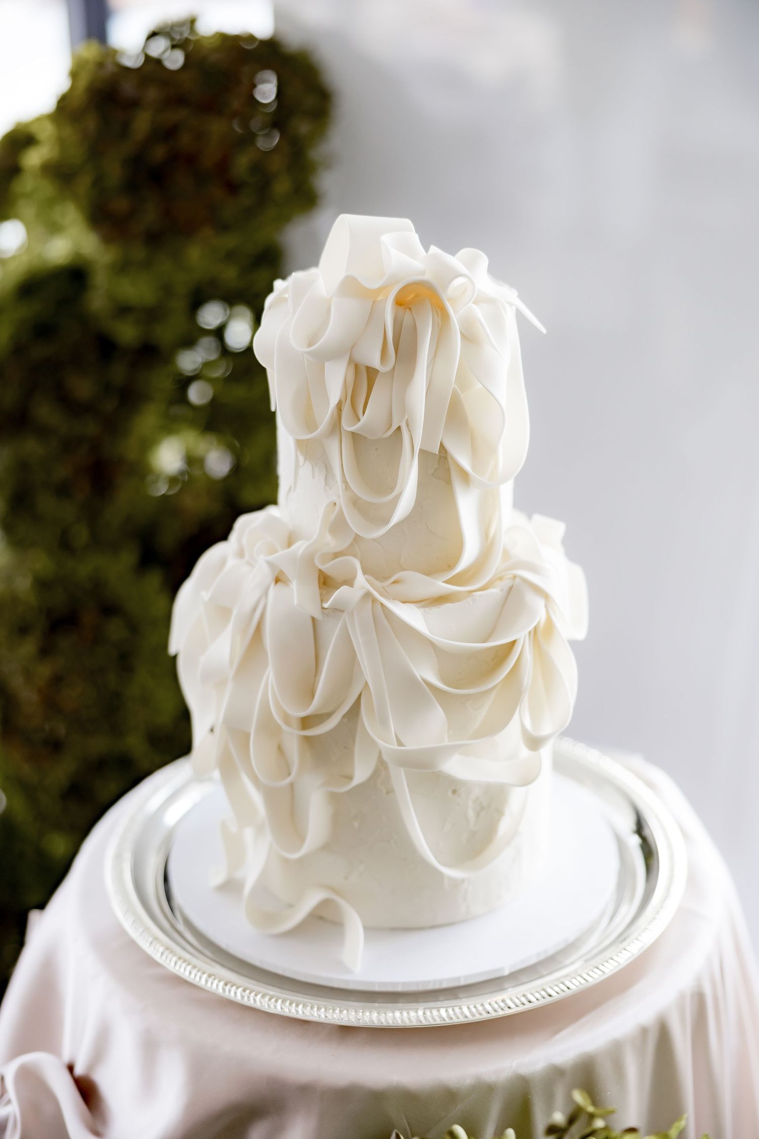Elegant white wedding cake with delicate ruffled fondant details displayed on a white plate against a blurred background.